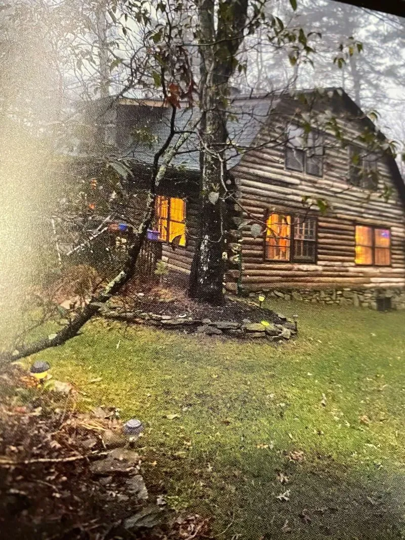 كوخ خشبي محاط بالأشجار، مضاء من الداخل، يمثل "كوخ الأمل" لعائلة روجرز في نورث كارولينا، قبل أن يتعرض للحرائق.