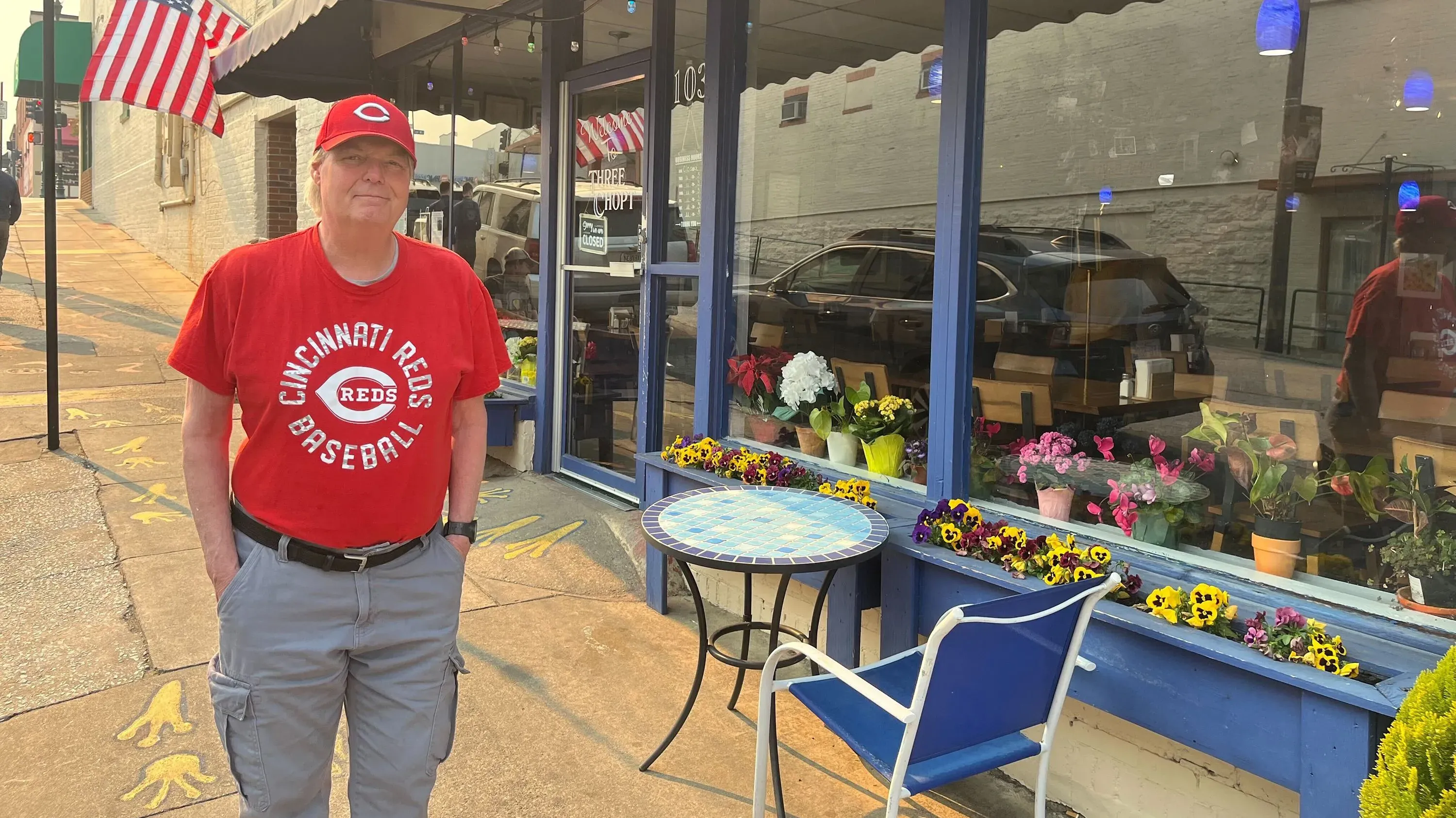 رجل يرتدي قميص فريق سينسيناتي ريدز يقف أمام مطعم مزين بالورود، مع لافتة أمريكية في الخلفية، يعكس روح التفاؤل بعد فقدان منزله.