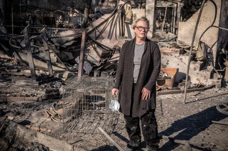 امرأة واقفة وسط أنقاض منزل محترق، تحمل حقيبة صغيرة، تعبيرها يعكس مشاعر الحزن والقلق بعد فقدان منزلها في حريق ألتادينا.