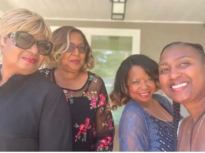 أربع نساء مبتسمات يتجمعن معًا في صورة سيلفي، يظهرن في أجواء غير رسمية، مع خلفية منزلية، تعكس روح الدعم والمودة.