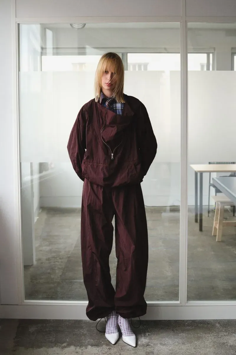 عارضة أزياء ترتدي بدلة فضفاضة باللون البني الداكن، وتقف في استوديو عصري شرق لندن، مع خلفية زجاجية.