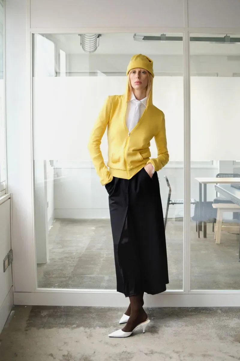 امرأة ترتدي سترة صفراء وقبعة، مع تنورة سوداء وأحذية بيضاء، تقف في استوديو عصري في شرق لندن، تعكس تصميمات الموضة البريطانية.