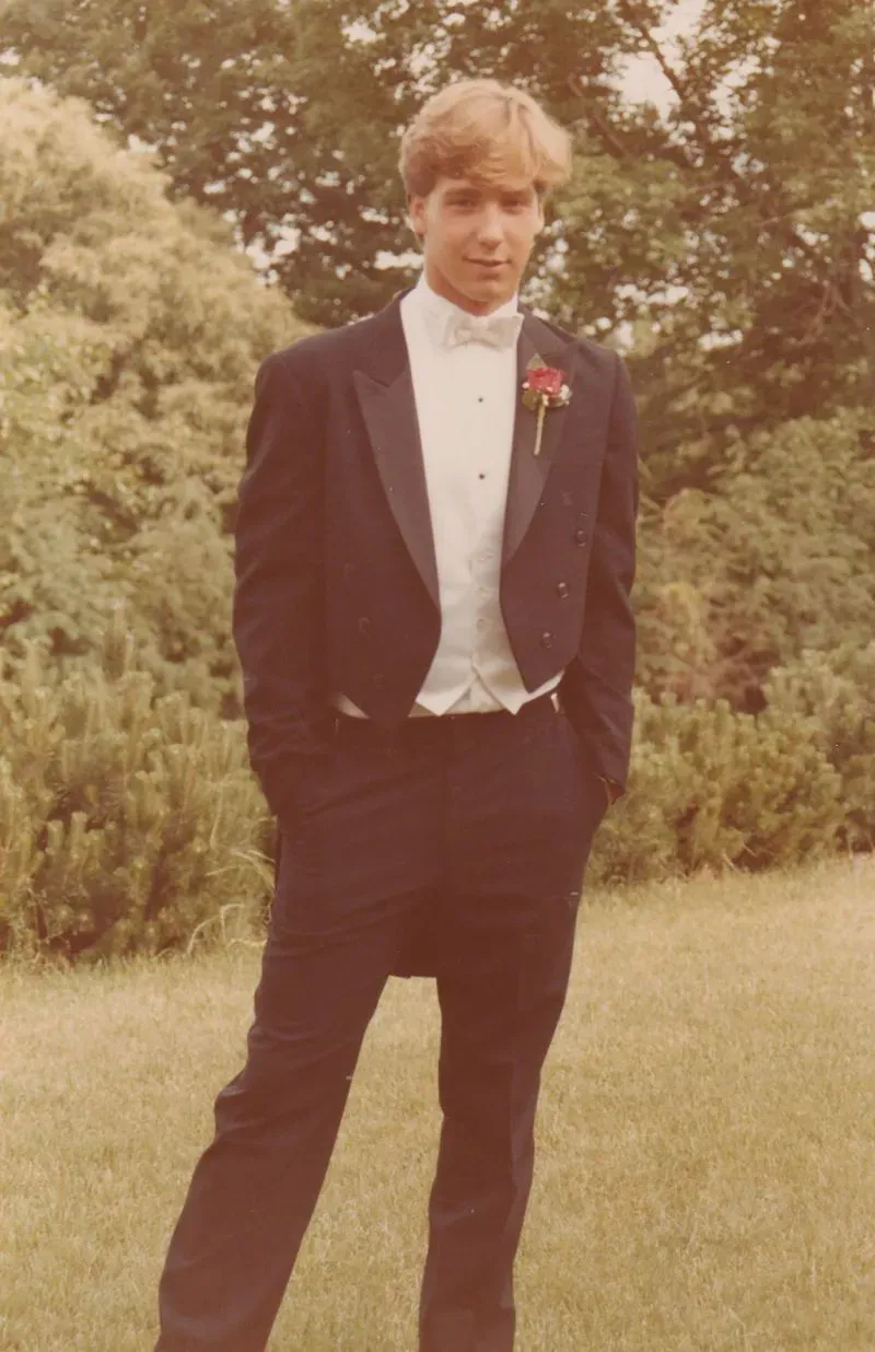 شاب يرتدي بدلة رسمية مع وردة على صدره، يقف في حديقة، يعكس لحظة من الحياة قبل مأساة تحطم طائرة بان آم 103.