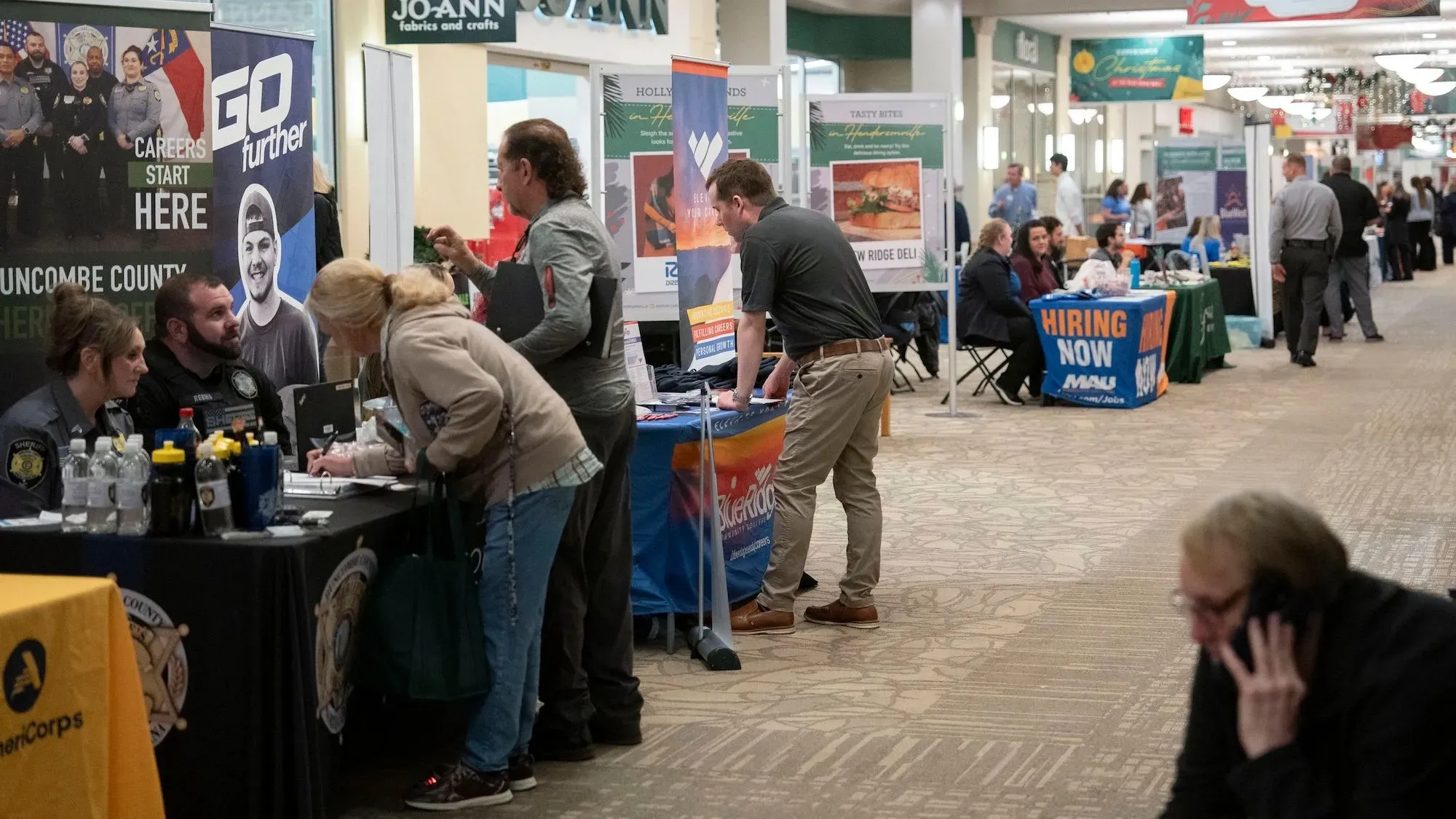 معرض توظيف مزدحم يظهر أشخاصًا يتحدثون مع ممثلين عن الشركات، مع لافتات تشير إلى وجود فرص عمل متاحة.