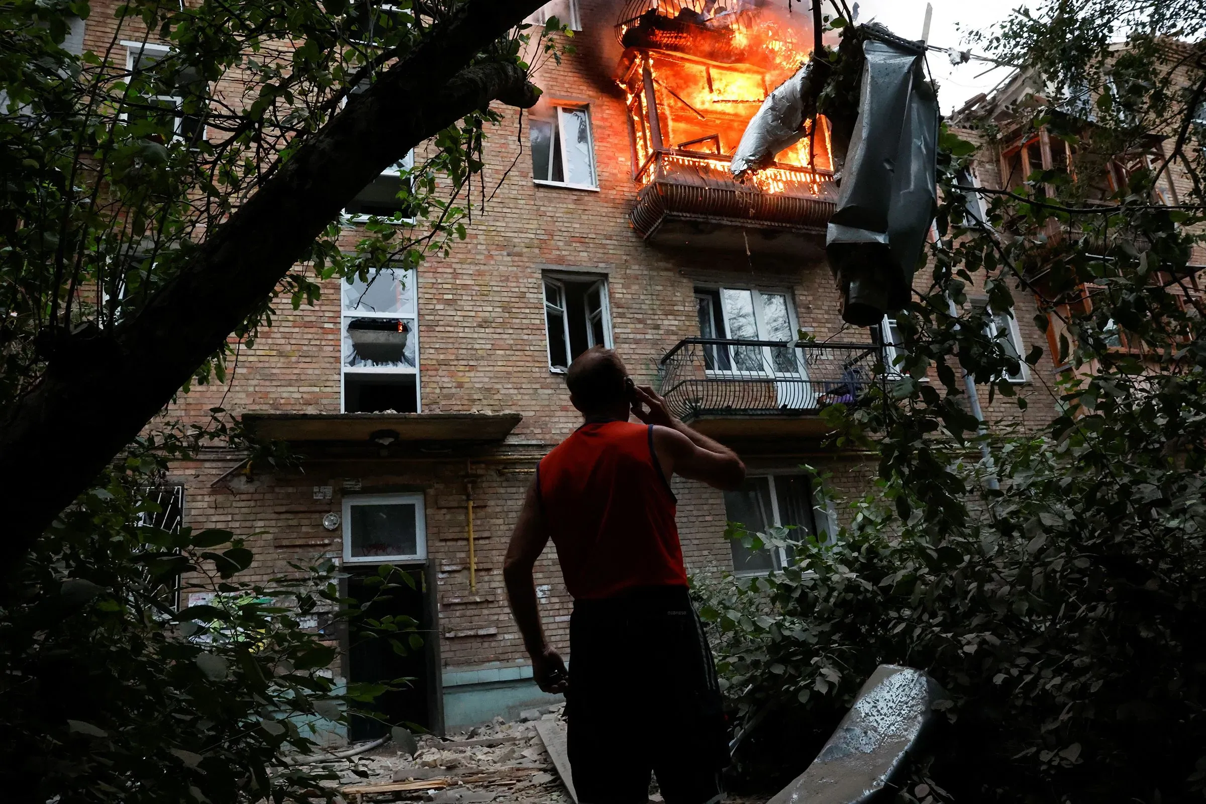 شخص يقف في حديقة أمام مبنى سكني محترق، حيث تتصاعد ألسنة اللهب من شرفة الطابق العلوي، مما يعكس آثار الصراع في أوكرانيا.
