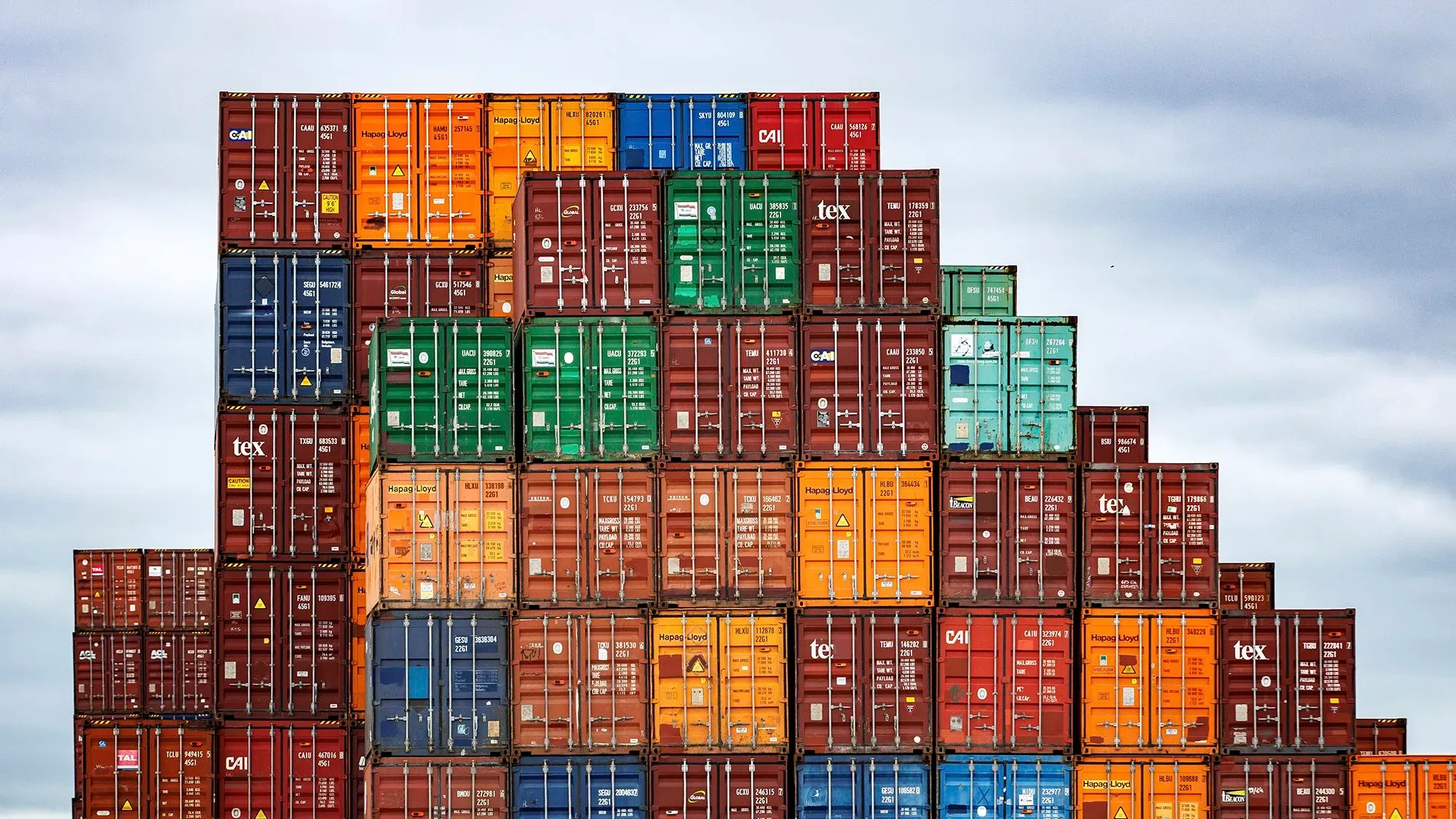 صناديق شحن ملونة متراكمة في ميناء، تعكس تأثير التعريفات الجمركية على التجارة الأمريكية والصناعات المحلية.