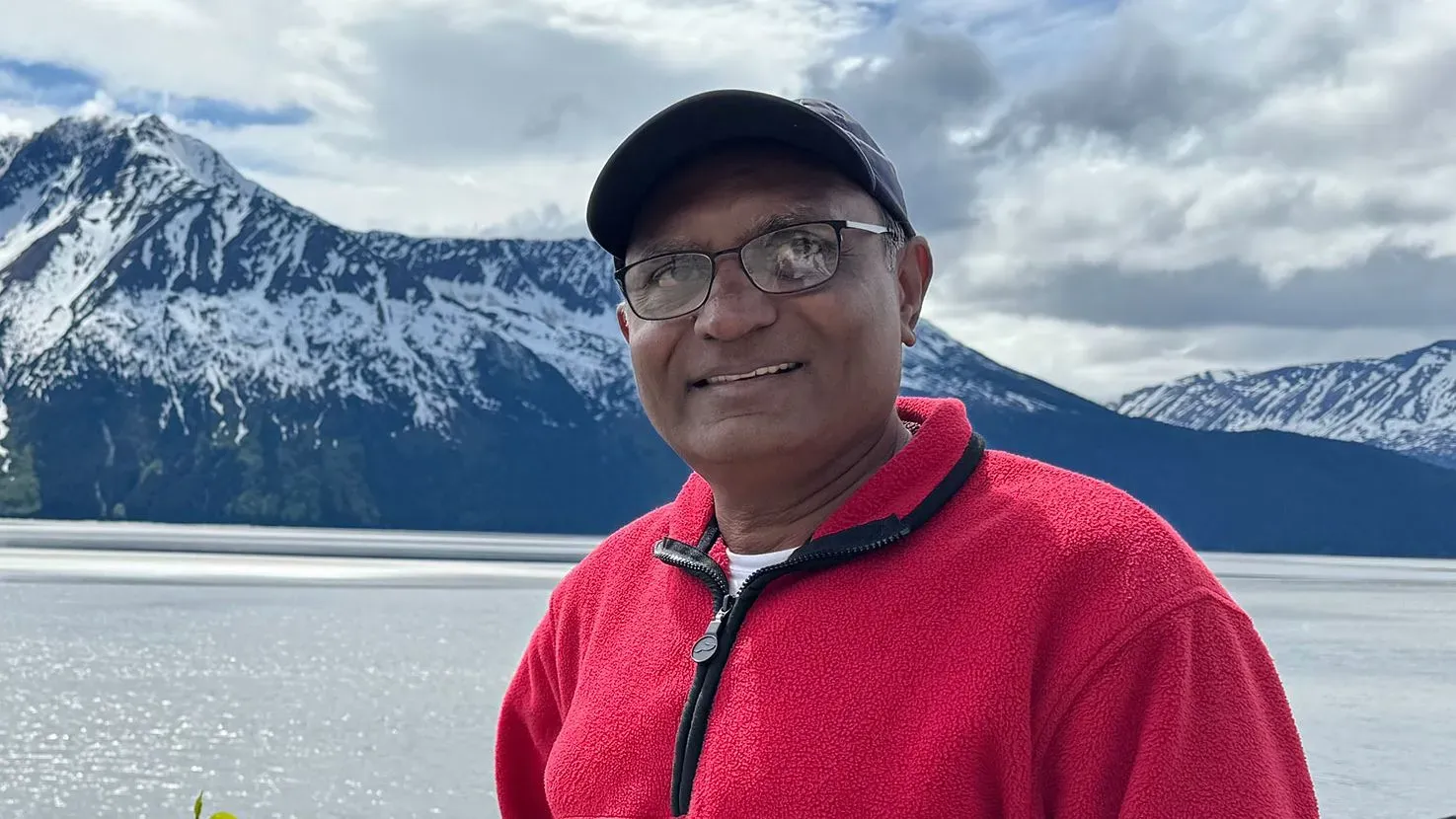 ماهيندرا باتيل مبتسم أمام منظر طبيعي جبلي، يرتدي سترة حمراء وقبعة، مع بحيرة خلفه تعكس السماء الملبدة بالغيوم.