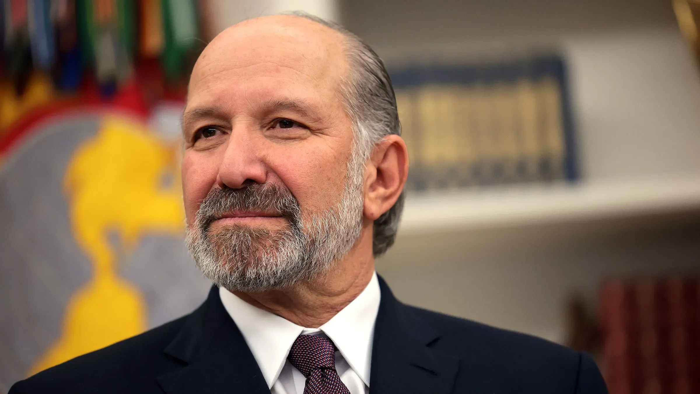 وزير التجارة هوارد لوتنيك، مبتسم، يتحدث في مؤتمر صحفي حول السياسات التجارية والاقتصادية لإدارة ترامب، مع خلفية رموز وطنية.