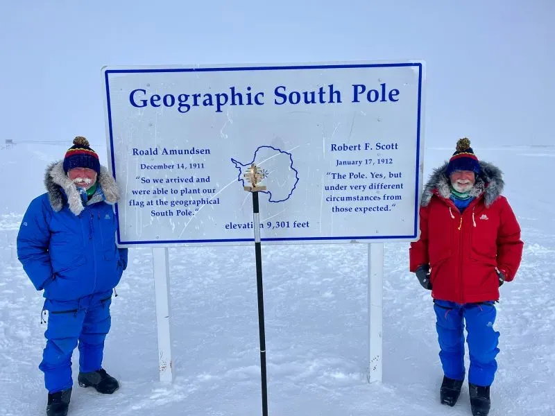 تشامبرز وزميله يقفان أمام لافتة القطب الجنوبي الجغرافي، مرتديين ملابس دافئة في بيئة ثلجية، مع التركيز على جهودهم العلمية لمكافحة التلوث البلاستيكي.