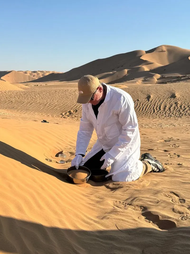 العالم آلان تشامبرز يجمع عينات من الرمال في الصحراء، مرتديًا معطفًا أبيض، في إطار بحثه عن تأثير اللدائن الدقيقة.