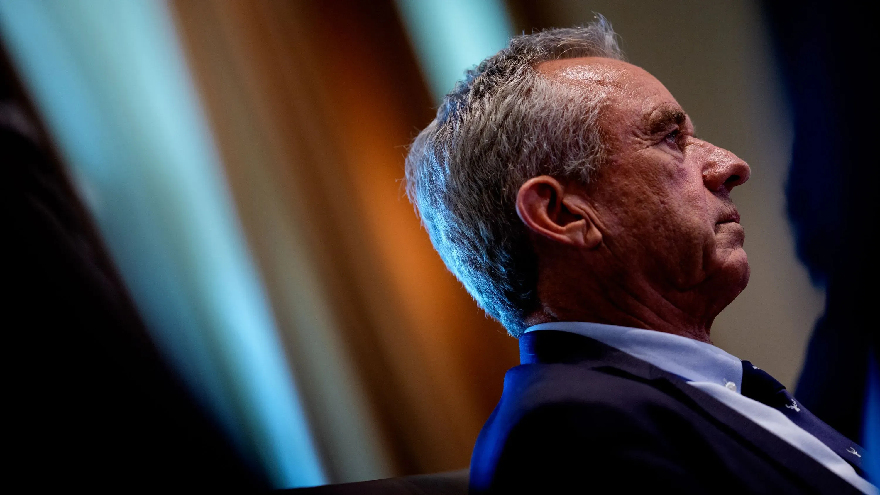 تظهر الصورة شخصًا يجلس في اجتماع، مع التركيز على تعبير وجهه الجاد. تتعلق الصورة بتحديثات لقاحات كوفيد-19 ومتطلبات السلامة الجديدة.