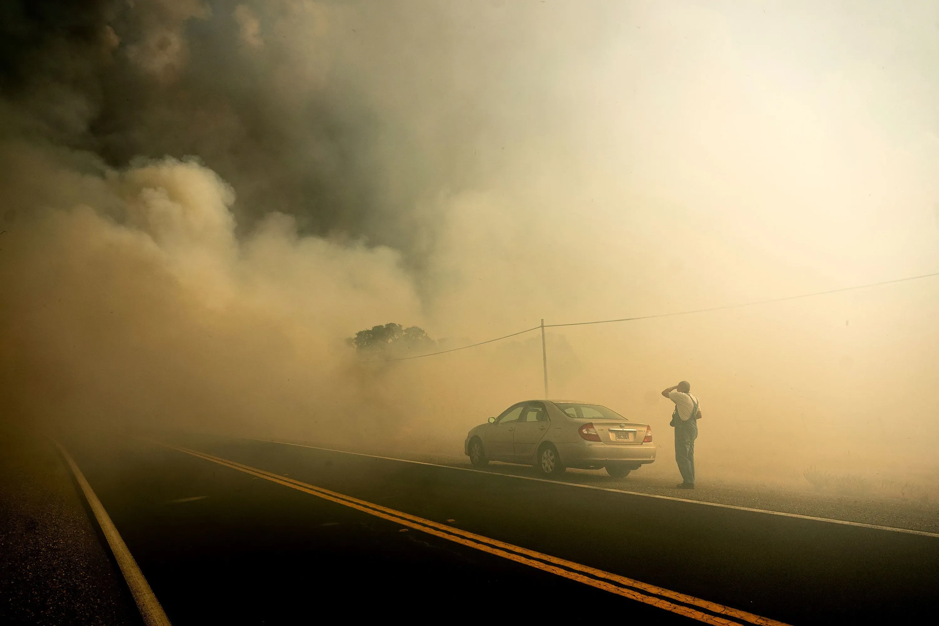 رجل يقف بجانب سيارته على طريق يعاني من كثافة الدخان الناتج عن حريق غابات في كاليفورنيا، وسط ظروف خطرة.
