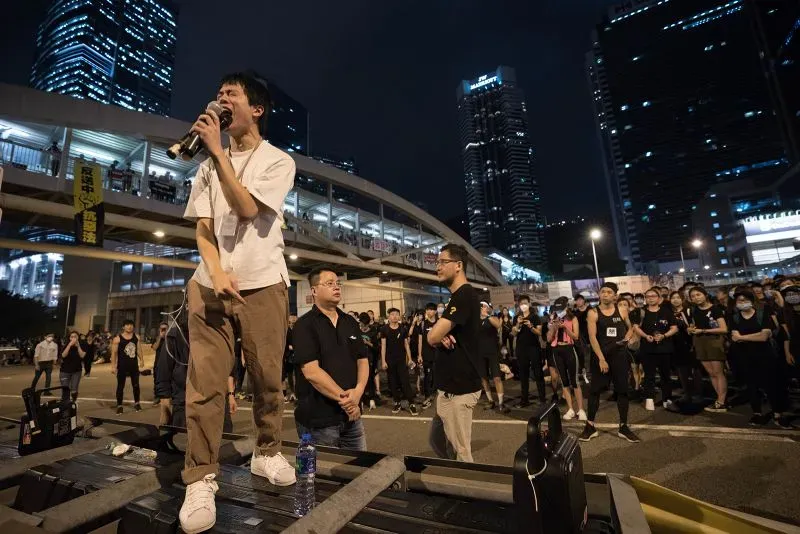 شخص يتحدث في تجمع حاشد في هونغ كونغ، مع خلفية لمباني شاهقة، يعكس روح الاحتجاج والدعوة للديمقراطية في المدينة.