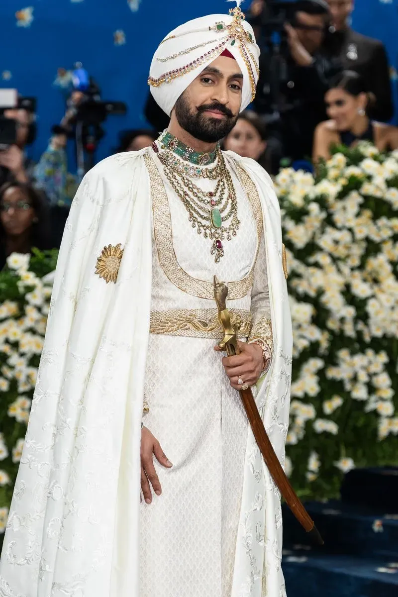 رجل يرتدي زيًا تقليديًا مزينًا بالأحجار الكريمة في حفل الميت غالا، مع خلفية من الزهور البيضاء، مما يعكس تأثير الموضة الكلاسيكية.