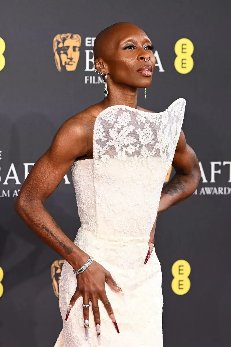 ممثلة ترتدي فستاناً أبيض أنيقاً في حفل جوائز البافتا، مع مجوهرات ماسية وزمردية، تعكس أناقة التصاميم الكلاسيكية.