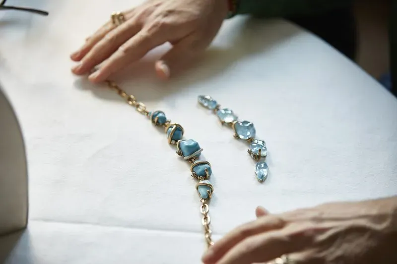 تصميم مجوهرات يتضمن قلادة وزخارف زرقاء، مع يدين تعملان على ترتيبها، تعكس الاتجاه المتزايد لاستخدام المجوهرات التراثية في الفعاليات الكبرى.