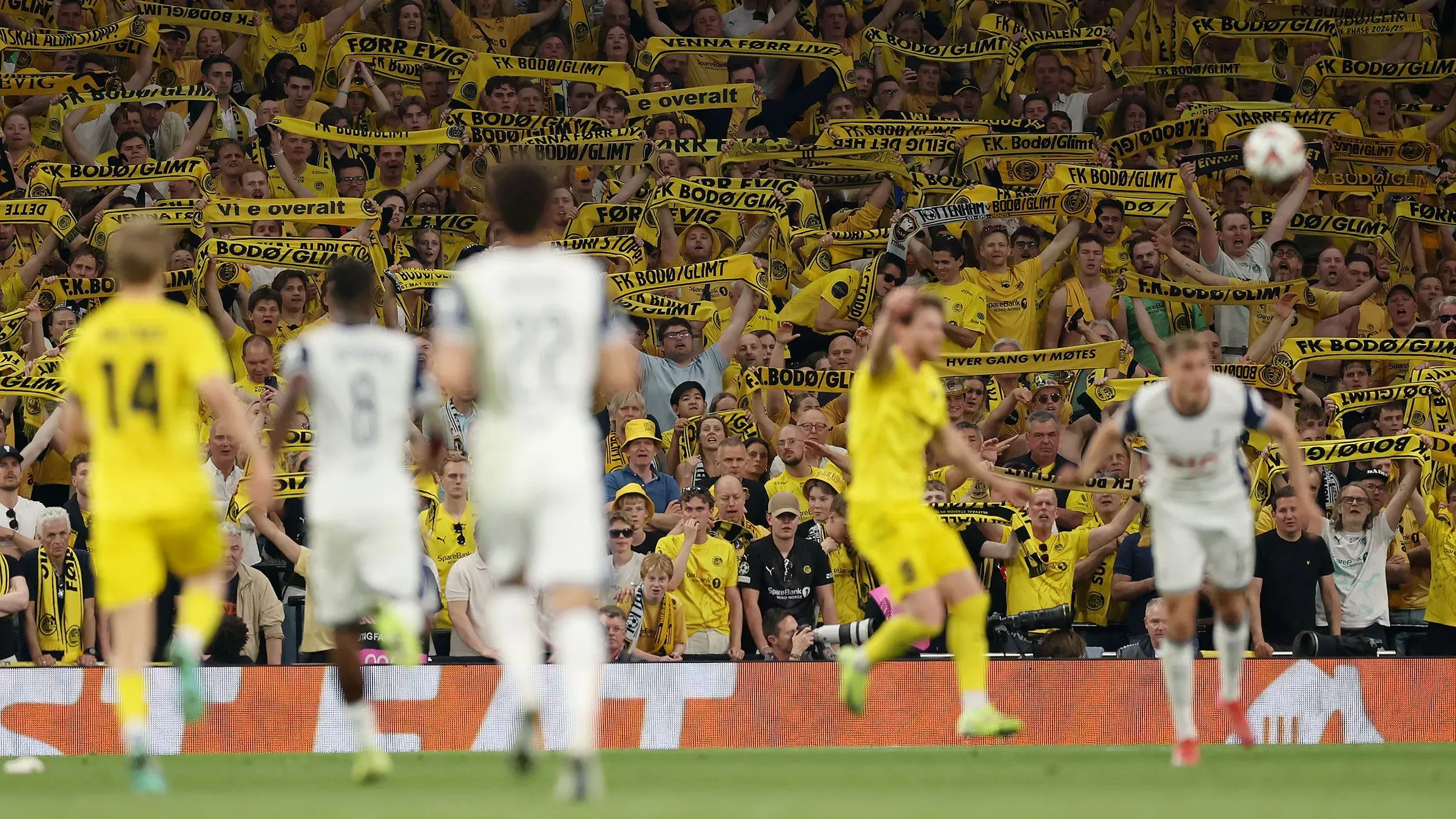 جمهور متحمس لفريق بودو/غليمت يحمل لافتات صفراء خلال مباراة نصف النهائي ضد توتنهام، مع التركيز على الأجواء الحماسية في الملعب.