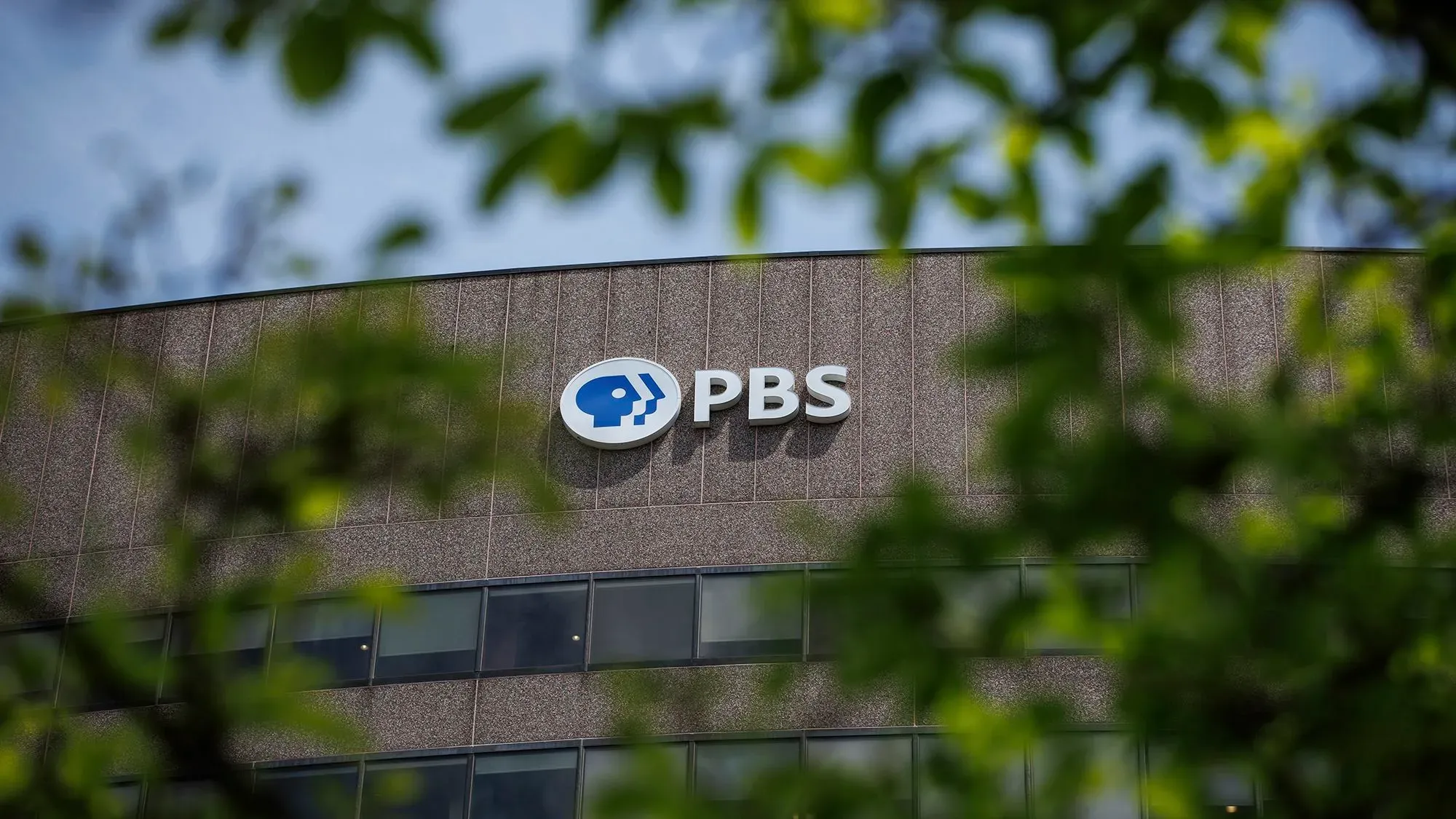 شعار شبكة PBS يظهر على واجهة المبنى، مع تركيز على أهمية التمويل الفيدرالي للمساعدات الإعلامية في ظل التخفيضات المقترحة.