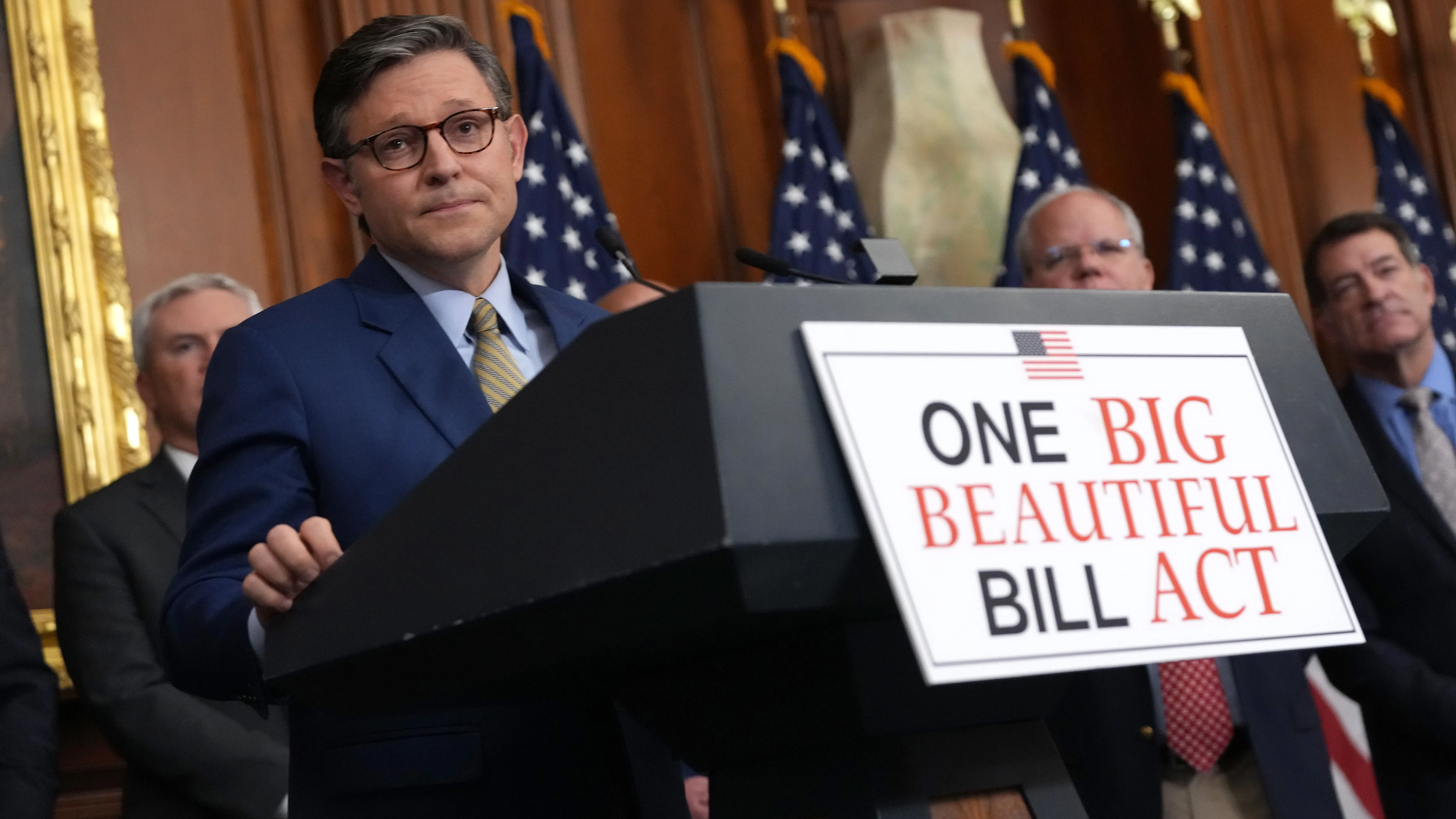 رجل يرتدي بدلة زرقاء يقف أمام منصة تحمل لافتة "مشروع قانون واحد كبير وجميل"، مع وجود مسؤولين آخرين خلفه، في مؤتمر صحفي حول التخفيضات الضريبية.