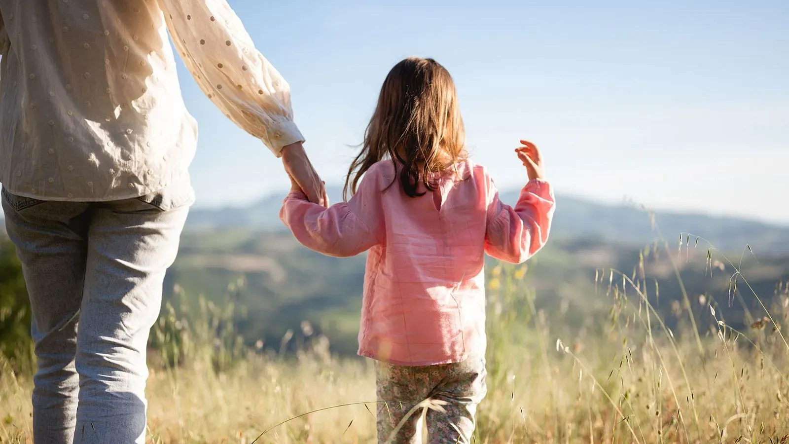 امرأة تمسك بيد طفلة صغيرة، يتجولان في منطقة طبيعية مع مناظر جبلية في الخلفية، مما يعكس الروابط الأسرية والهدوء.