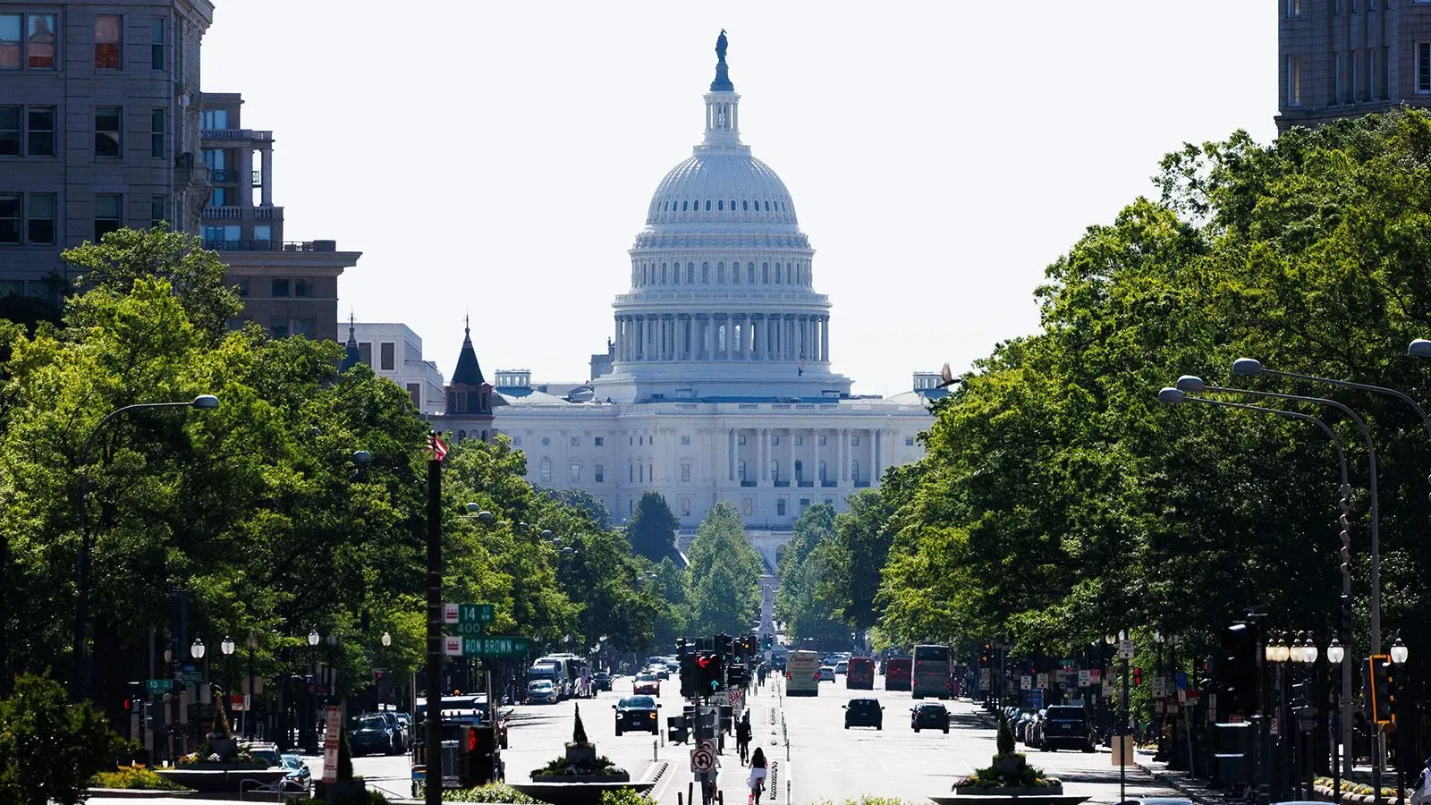 مبنى الكابيتول الأمريكي يظهر في الخلفية، محاطاً بالأشجار، مع طريق مزدحم أمامه، مما يعكس النشاط السياسي في واشنطن.