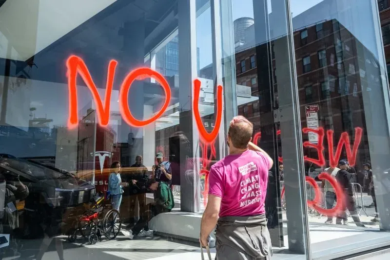 شخص يقوم بتنظيف جدار زجاجي لمتجر تسلا، حيث تظهر عليه كتابات حمراء تعبر عن احتجاجات ضد إيلون ماسك، مع تجمع عدد من المارة.