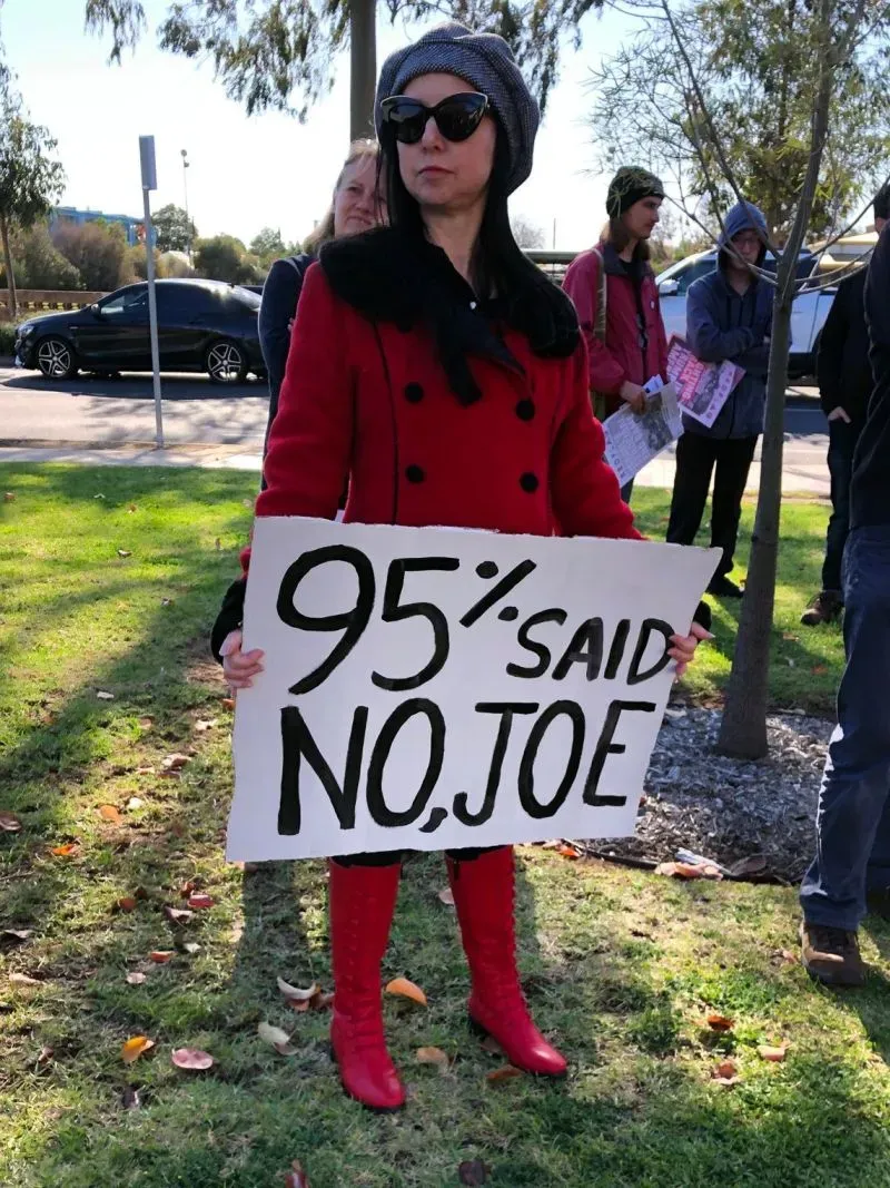 امرأة ترتدي معطفًا أحمر وقبعة، تحمل لافتة مكتوب عليها "95% قالوا لا، جو". تظهر في احتجاج ضد مصنع تسلا لإعادة تدوير البطاريات في تونزلي، أستراليا.