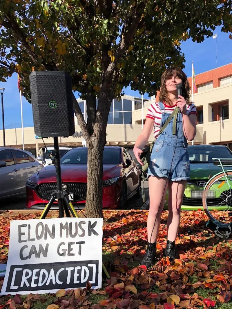 امرأة تتحدث في تجمع احتجاجي تحت شجرة، تحمل ميكروفونًا، بجوار لافتة مكتوب عليها "إيلون ماسك يمكن أن يحصل على [مُحذَف]"، مع سيارات في الخلفية.