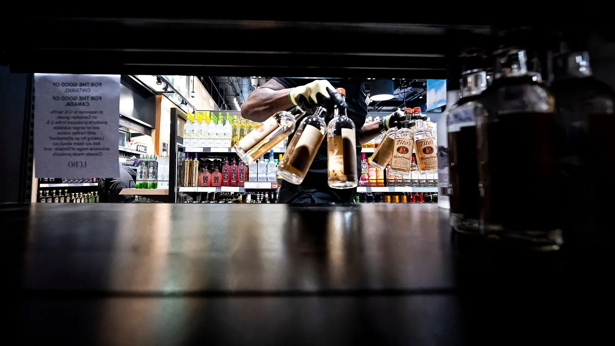 يد تظهر شخصًا يرتدي قفازات وهو يحمل زجاجات مشروبات كحولية من رف في متجر، مع وجود لافتة توضح تأثير الرسوم الجمركية على الواردات الأمريكية.