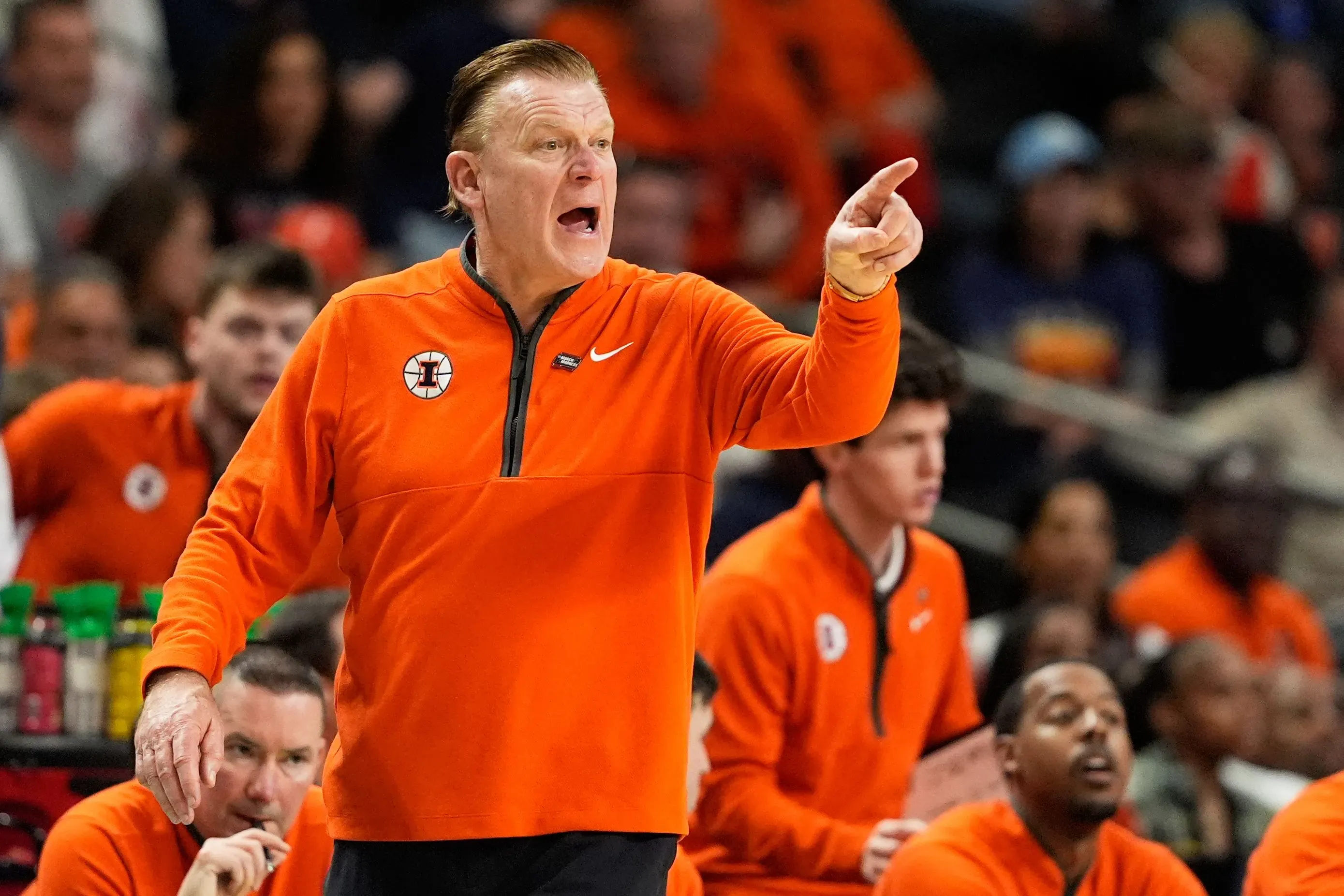 مدرب كرة السلة براد أندروود يتحدث بحماس خلال مباراة، مرتديًا سترة برتقالية، مع لاعبي فريق إلينوي خلفه.