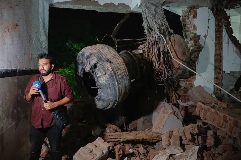 مراسل يقف أمام حطام طائرة بوينج 787 دريملاينر في موقع الحادث، مع وجود أجزاء من المباني المدمرة حوله.