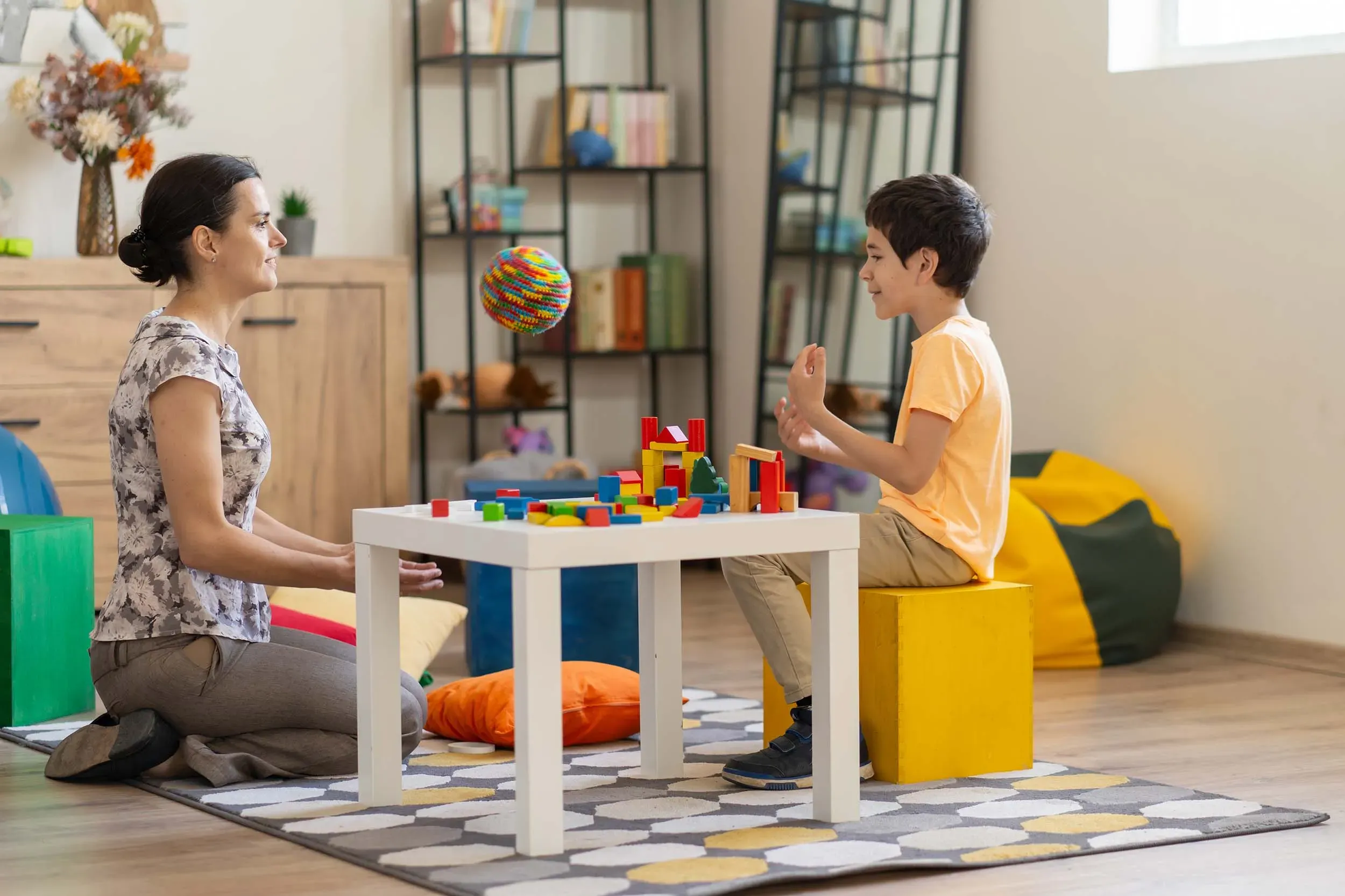 امرأة وطفل يلعبان معًا على طاولة، حيث يقوم الطفل ببناء هياكل باستخدام مكعبات ملونة، مما يعكس تفاعلًا إيجابيًا وداعمًا.