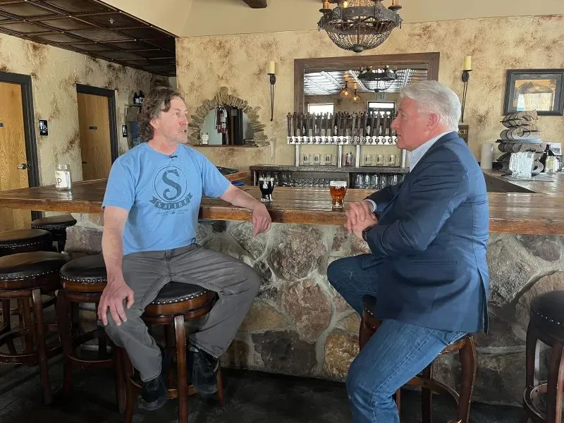 محادثة بين رجلين في بار بمزرعة كولورادو، يتبادلان الآراء حول السياسة والزراعة، مع خلفية من الأثاث الخشبي والمشروبات.