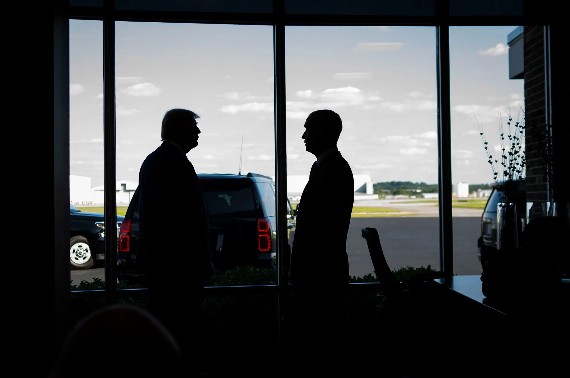 صورة تظهر ظلال كريستي نويم وكوري ليفاندوفسكي في مطار، مع خلفية تضم سيارات ومشهد السماء، تعكس تأثير ليفاندوفسكي في وزارة الأمن الداخلي.