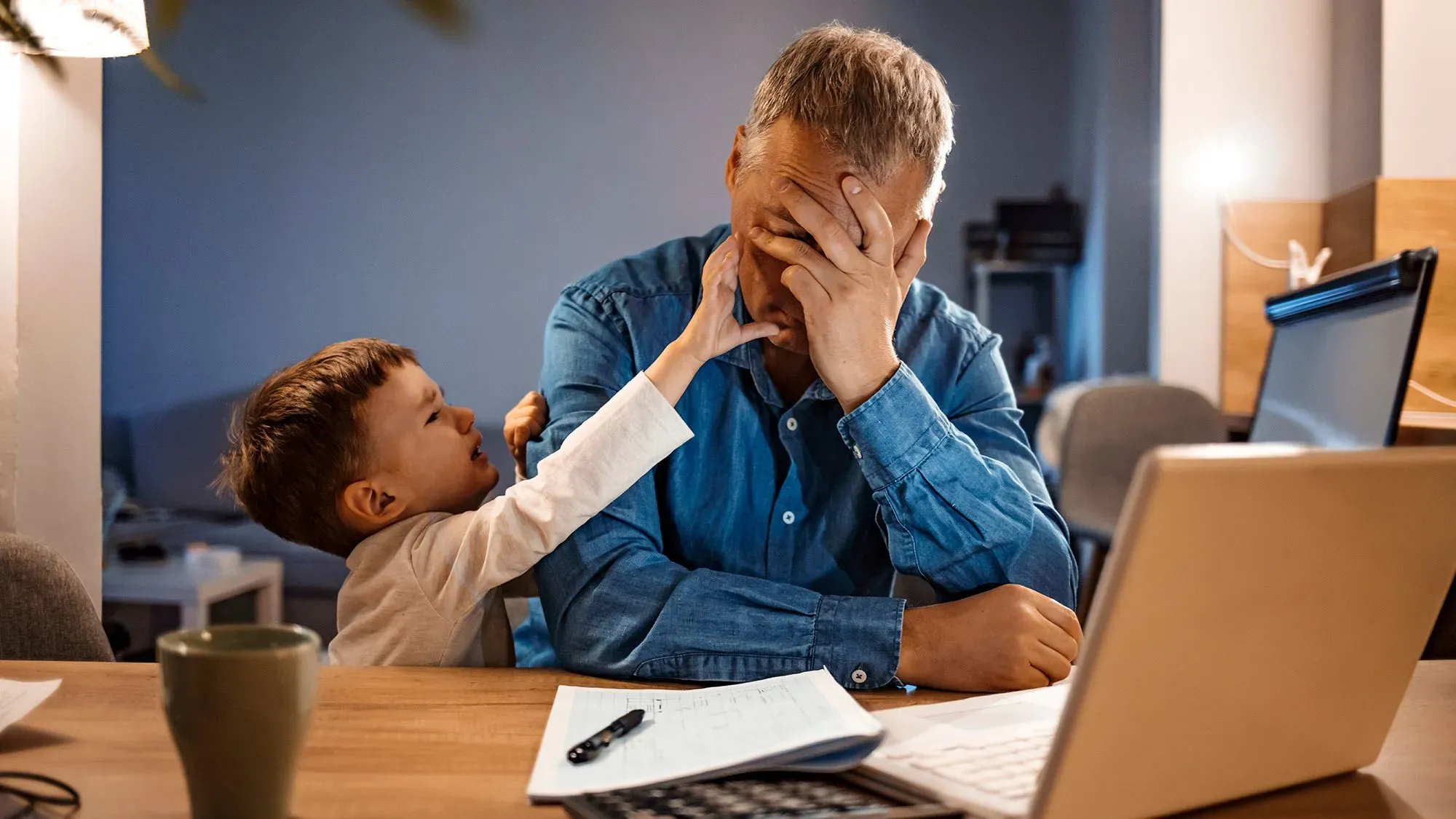 رجل في منتصف العمر يجلس أمام حاسوب، يبدو متوترًا ويضع يده على وجهه، بينما يحاول طفله الصغير لفت انتباهه.