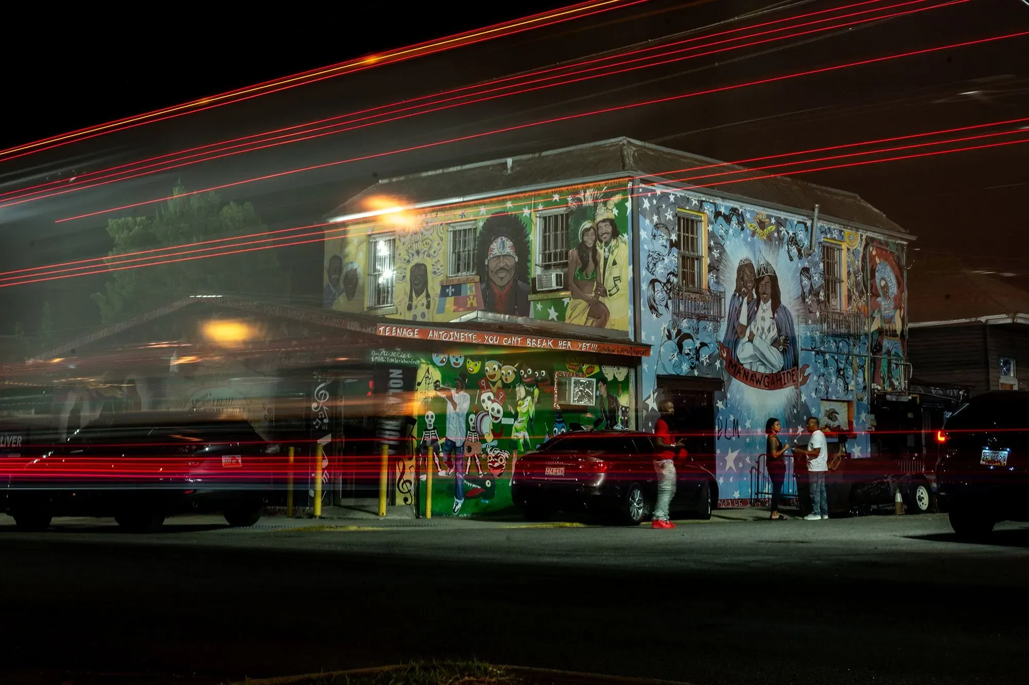 مبنى مزين برسوم جدارية ملونة في نيو أورلينز، مع سيارات متوقفة وأشخاص يتحدثون في الشارع، مما يعكس الثقافة المحلية النابضة بالحياة.