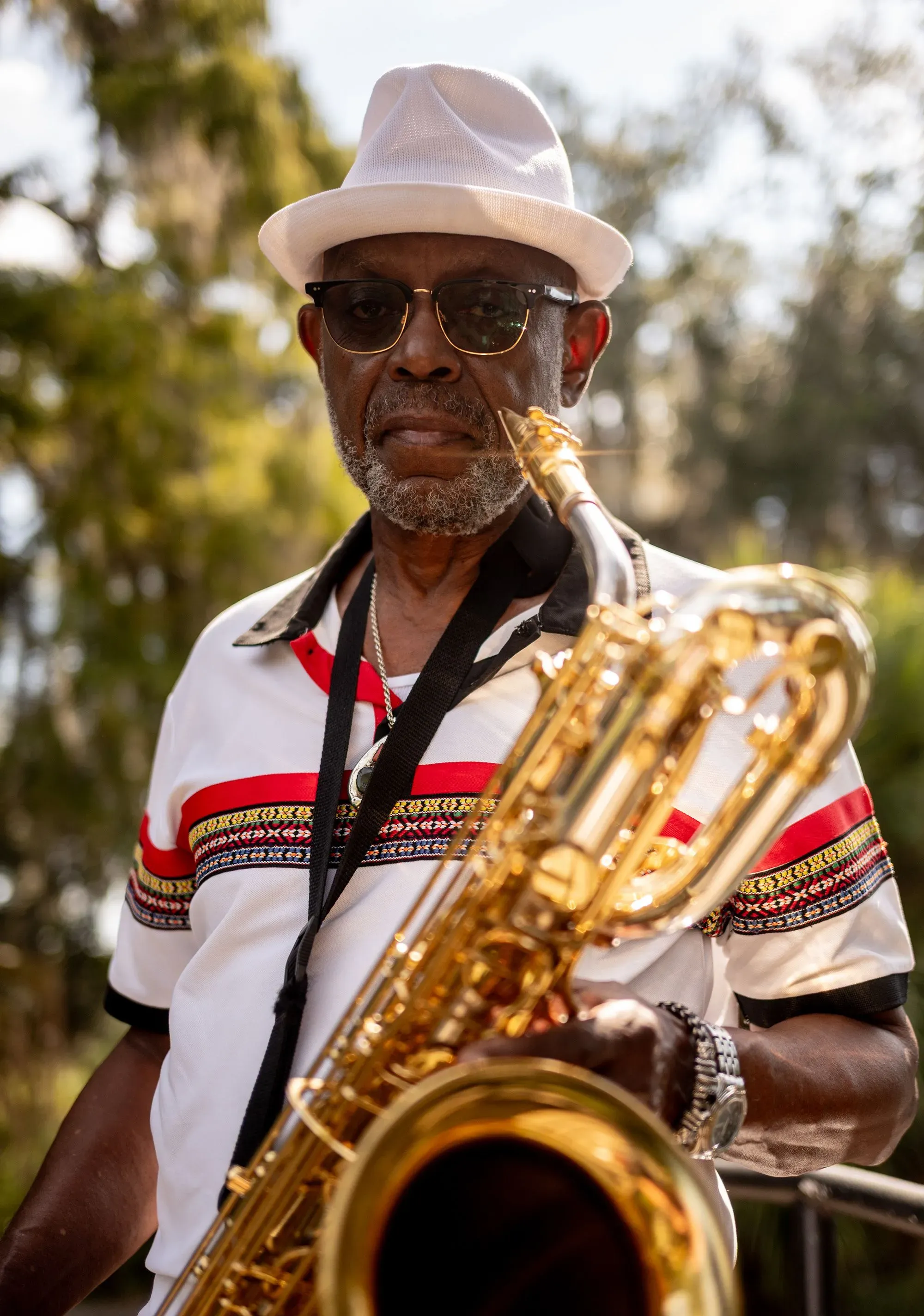 رجل مسن يرتدي نظارات وقميصًا مزخرفًا، يحمل آلة الساكسفون، مع خلفية طبيعية تُظهر أشجارًا خضراء، تعكس روح الموسيقى في نيو أورلينز.