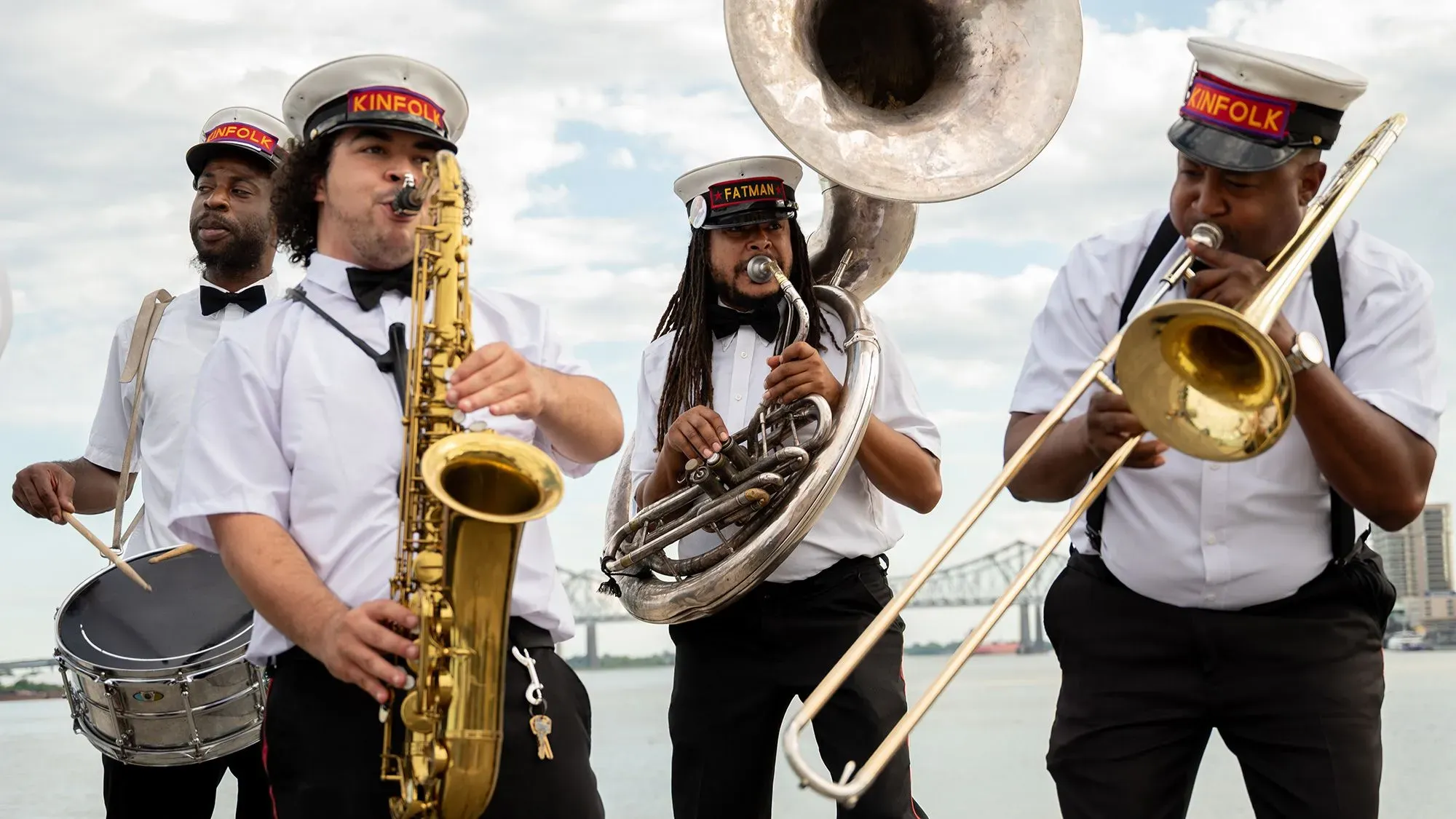 فرقة نحاسية تؤدي في نيو أورلينز، تضم عازفين على الساكسفون والترومبون والطبول، مع خلفية نهرية، تعكس الثقافة الموسيقية الغنية للمدينة.