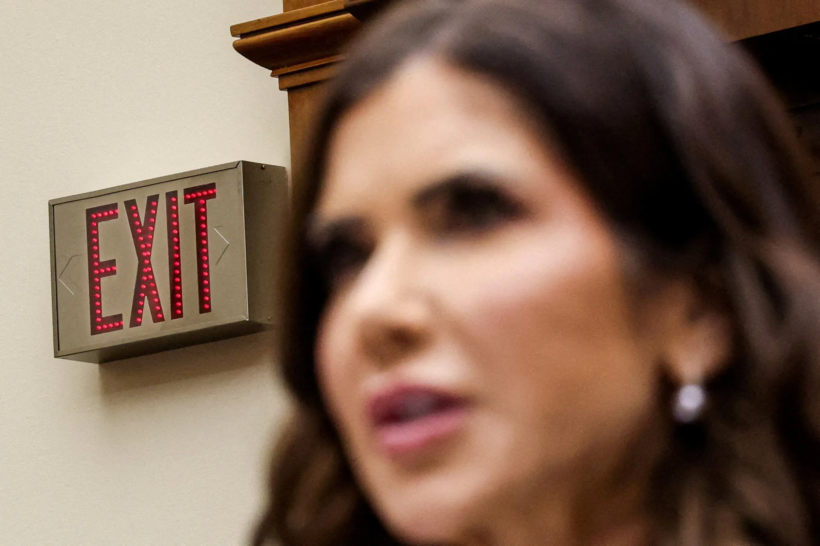 لافتة "خروج" مضيئة في خلفية الصورة مع تركيز على وجه كريستي نويم أثناء جلسة استماع في الكونغرس، تعكس التوترات السياسية حول إقالتها.