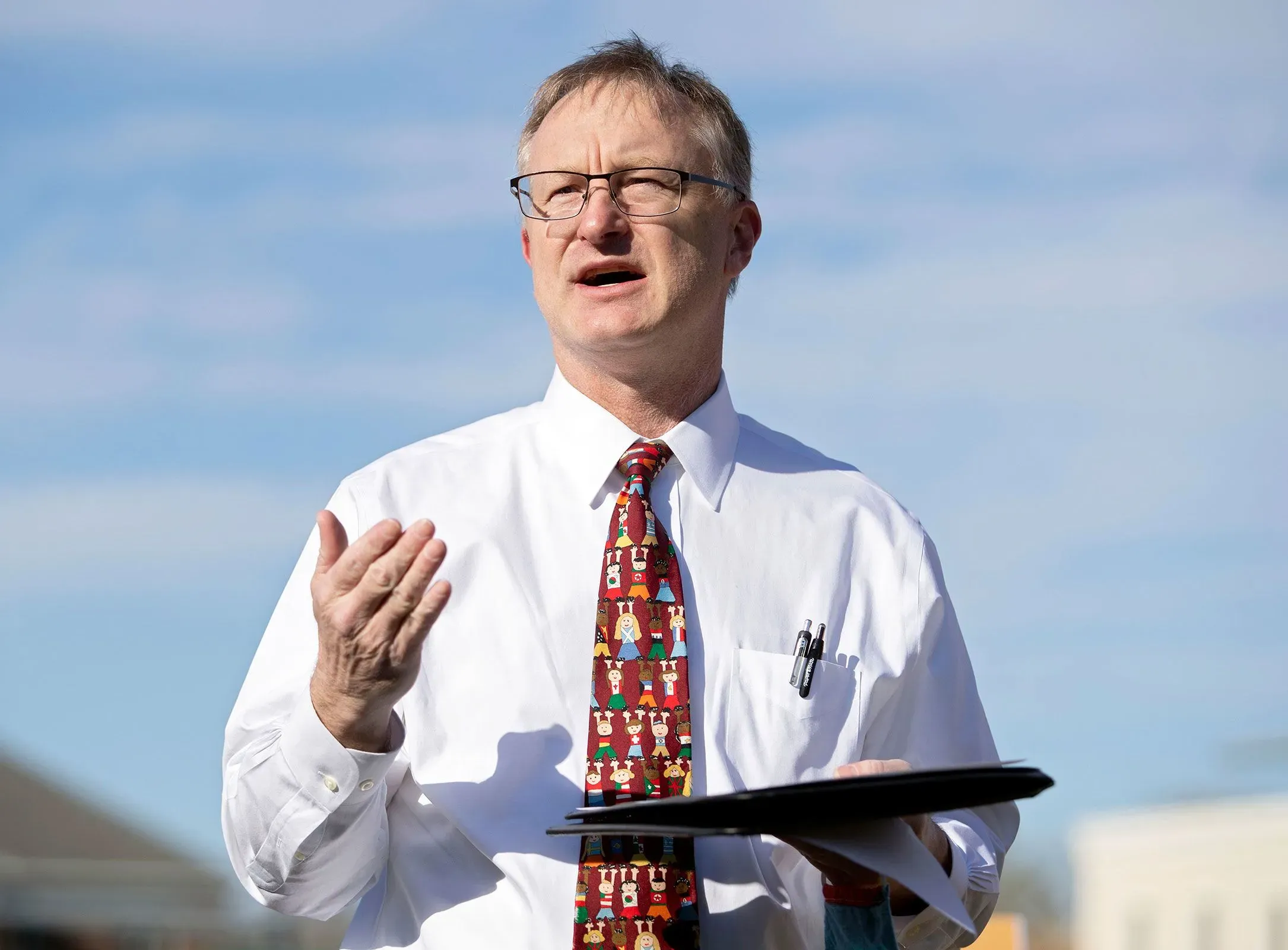 رجل يرتدي قميصًا أبيض وربطة عنق ملونة، يتحدث بجدية في الهواء الطلق، مع سماء زرقاء في الخلفية، يعبر عن قضايا الرعاية الصحية في تشيرشفيل.