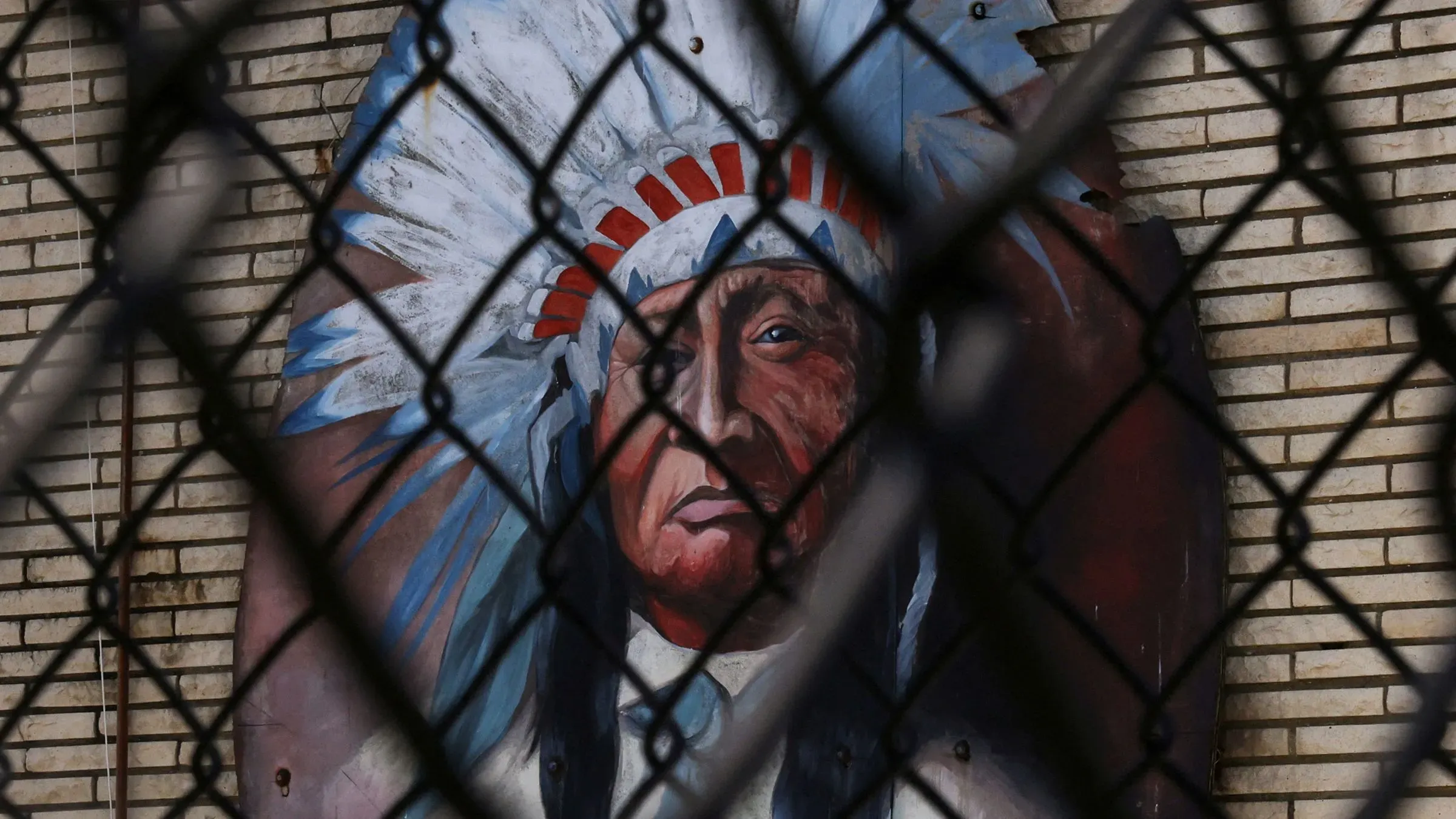 صورة لجدارية تُظهر وجه هندي أمريكي يرتدي غطاء رأس مزخرف، محاطة بسياج معدني، تعكس الجدل حول تمائم الفرق الرياضية.