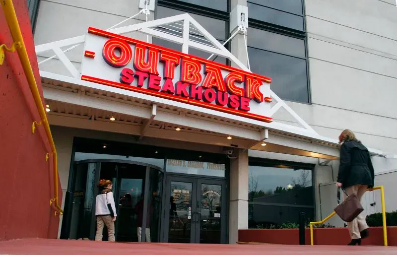 واجهة مطعم آوتباك ستاكس، مع لافتة مضيئة تحمل اسم المطعم. تظهر امرأة تدخل المطعم، مما يعكس تراجع شعبية آوتباك وسط المنافسة.