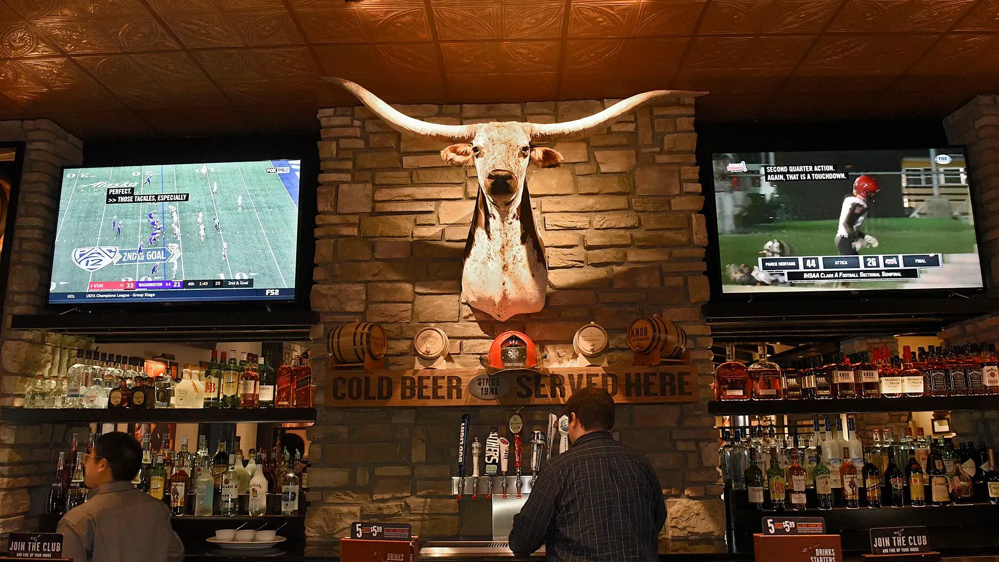 ديكور مطعم يبرز رأس بقرة على الجدار، مع شاشات تعرض المباريات، وزبون يتفاعل مع البار، مما يعكس أجواء تكساس رودهاوس المفعمة بالحيوية.