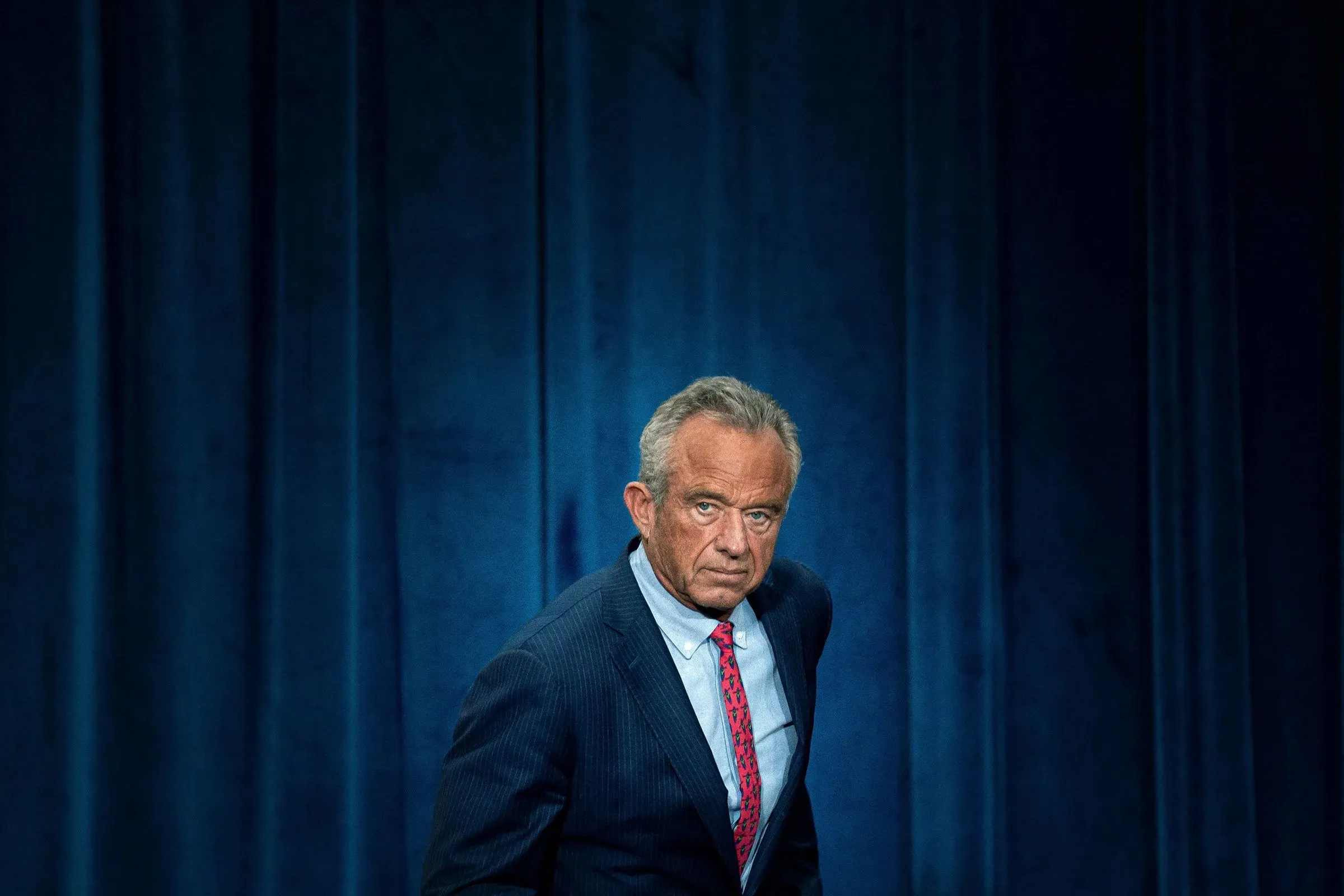 روبرت كينيدي جونيور، وزير الصحة والخدمات الإنسانية، يبدو جادًا أثناء حديثه في مؤتمر، مع خلفية زرقاء.