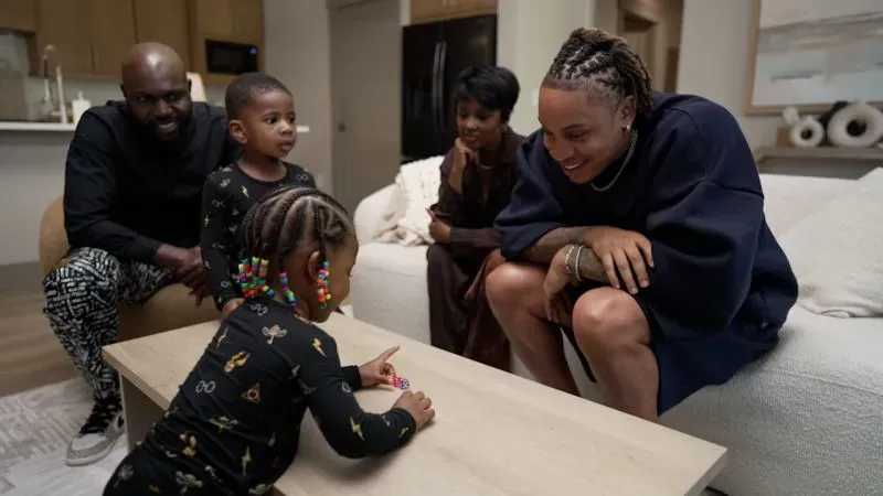 عائلة مختلطة الثقافات تتفاعل في غرفة معيشة دافئة، حيث يلعب طفل على الطاولة بينما يراقبه البالغون بابتسامات محبة.