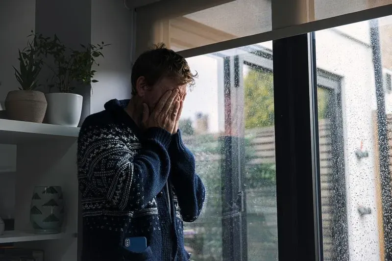 رجل يرتدي سترة داكنة، يغطي وجهه بيديه، ويبدو متوترًا أمام نافذة مبللة بالمطر، مما يعكس مشاعر القلق والإجهاد.