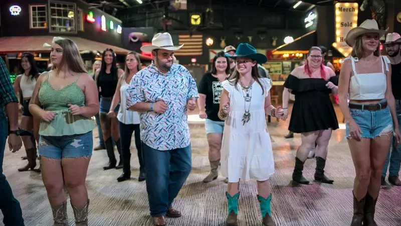 مجموعة من الأشخاص يرتدون قبعات رعاة البقر ويرقصون في قاعة رقص، مما يعكس أجواء اجتماعية نابضة بالحياة.