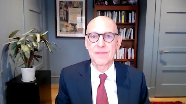 صورة لرجل يرتدي بدلة رسمية ونظارات، يجلس في مكتب مع خلفية تحتوي على كتب ونباتات، يتحدث عن قضايا التمويل الطبي.