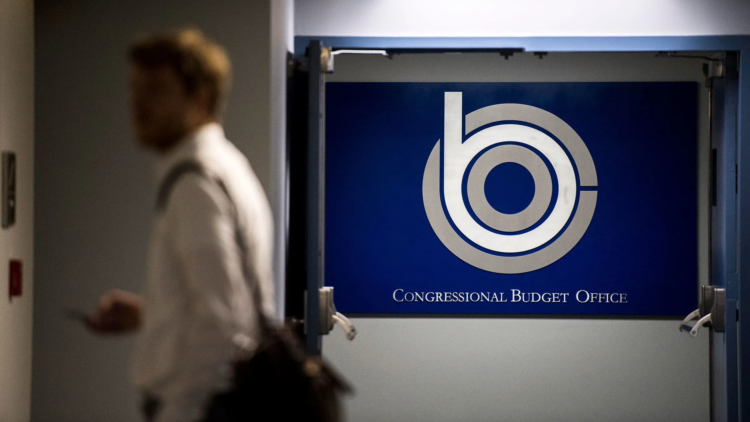 مكتب الميزانية في الكونغرس يظهر في الصورة، مع شخص يمشي بجانبه، مما يعكس الجدل حول تأثير التشريعات على العجز الوطني.