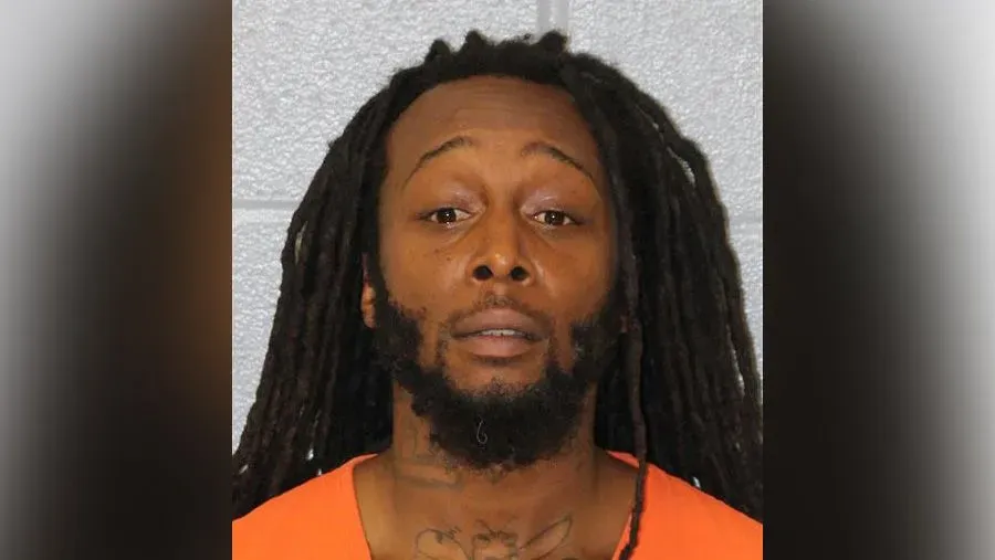 صورة لرجل ذو شعر مجعد طويل يرتدي سترة برتقالية، يظهر تعبيره الجاد. تم اعتقاله بتهمة القتل في حادثة طعن.